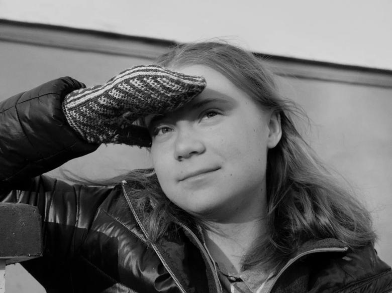 Black and white portrait of activist Greta Thunberg. She sits in a puffer jacket shielding her eyes from the sun in knitted mittens and is smiling slightly at the camera. 