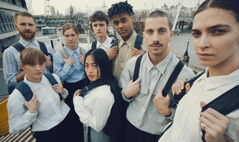 eight people stand on a staircase overlooking the southbank centre, they're all wearing white shirts and holding black backpack straps across their chests.