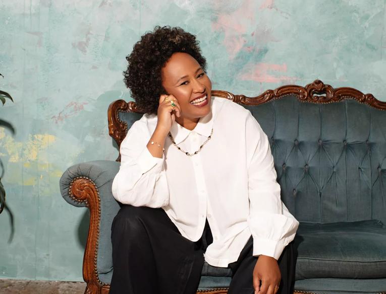 Singer Emeli Sandé sitting on a sofa with her head leaning against her hand and smiling. She wears black trousers, a white blouse and black loafers.