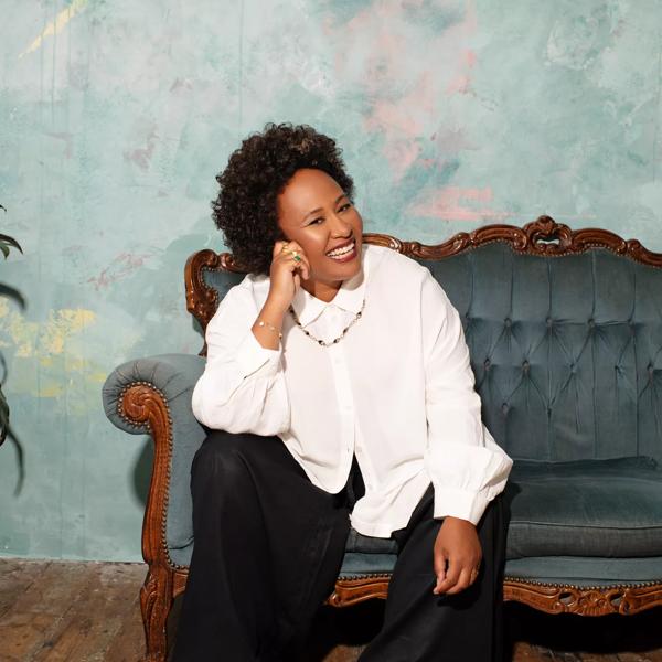 Singer Emeli Sandé sitting on a sofa with her head leaning against her hand and smiling. She wears black trousers, a white blouse and black loafers.