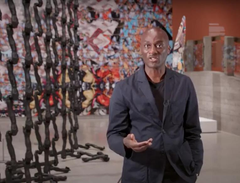 Curator Ekow Eshun talks to the camera inside Hayward Gallery exhibition, In the Black Fantastic. In the background we can see works by the artist Nick Cave.