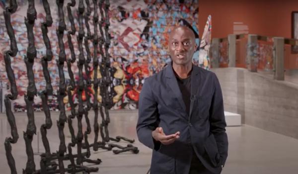 Curator Ekow Eshun talks to the camera inside Hayward Gallery exhibition, In the Black Fantastic. In the background we can see works by the artist Nick Cave.