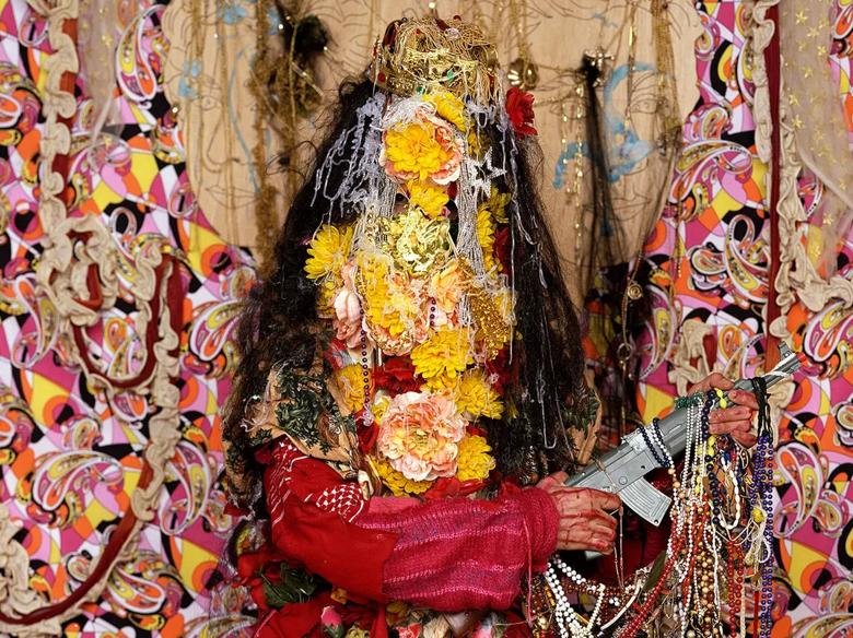 Hew Locke: Bounty killer, 2007 artwork of a person holding a machine gun, beeds and covered in flowers. Pink patterned backdrop behind the person