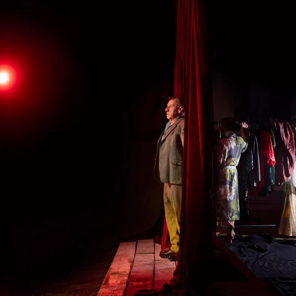 A man stands on stage against a red curtain, behind the curtain a woman stands holding a rail of clothing.