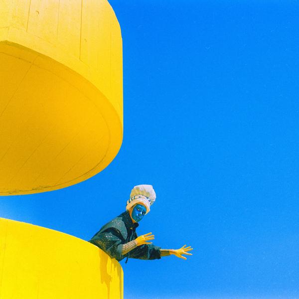 Artistic shot of a blue painted face person leaning over a yellow staircase over the blue sky