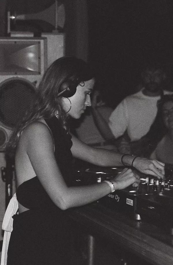 Black and white photo of a woman with long hair standing at DJ decks with headphones on.