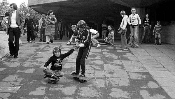 Young skateboarders in action at the undercroft skate space in 1976; in the foreground one skateboarder pulls along a nother one who is sitting on his board.