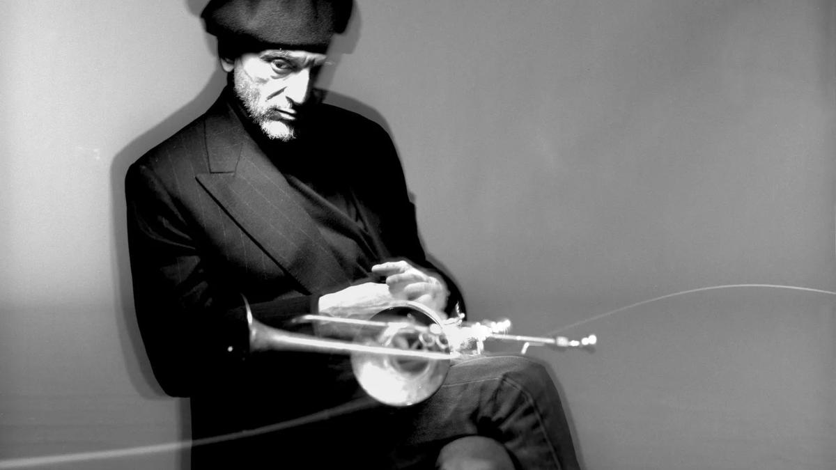 A black and white image of Tomasz Stanko sitting on a chair with his trumpet 