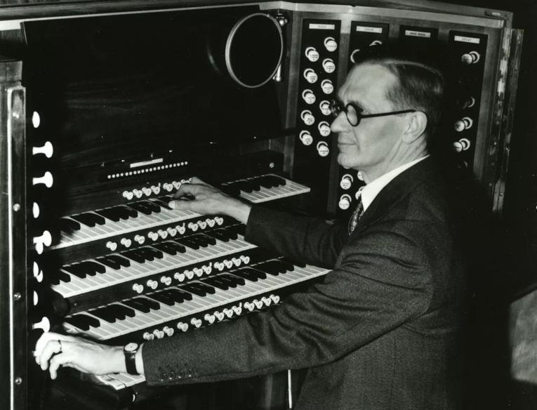 Ralph Downes, a White man wearing glasses and a suit, sits at the console, with its many keys and stops visible, of the Royal Festival Hall organ in an archive image believed to be from the 1950s