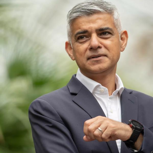 Sadiq Khan wears a charcoal suit with an open collar white shirt and is pictured against a blurred botanical background. He wears a black watch on his left wrist and looks up smiling off camera. 