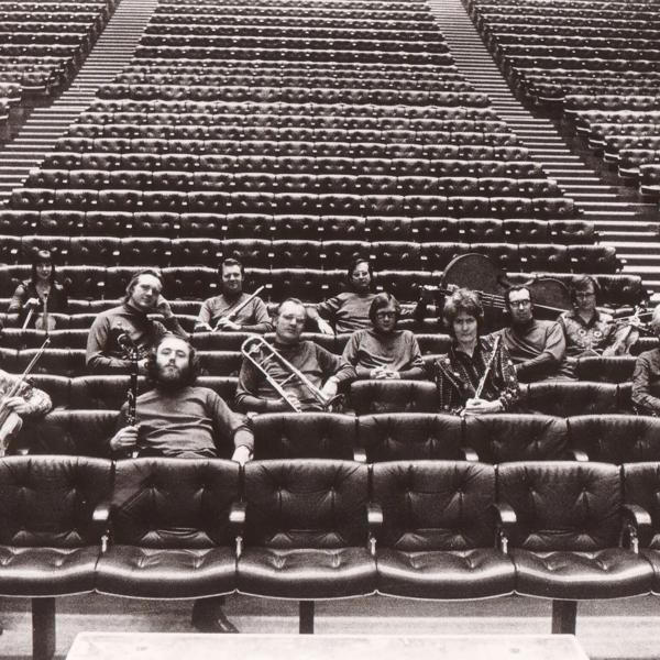 Black and white archive shot of 14 musicians and their instruments sitting in an otherwise empty concert hall