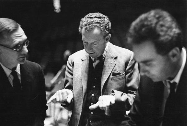 Ralph Downes and Benjamin Britten are in discussion; both men are wearing suits. An unknown third man can be seen closer to the lens