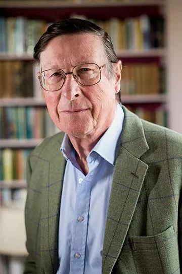 Portrait of Max Hastings, standing in front of full bookshelves. He wears glasses, a green tweed jacket and blue shirt 