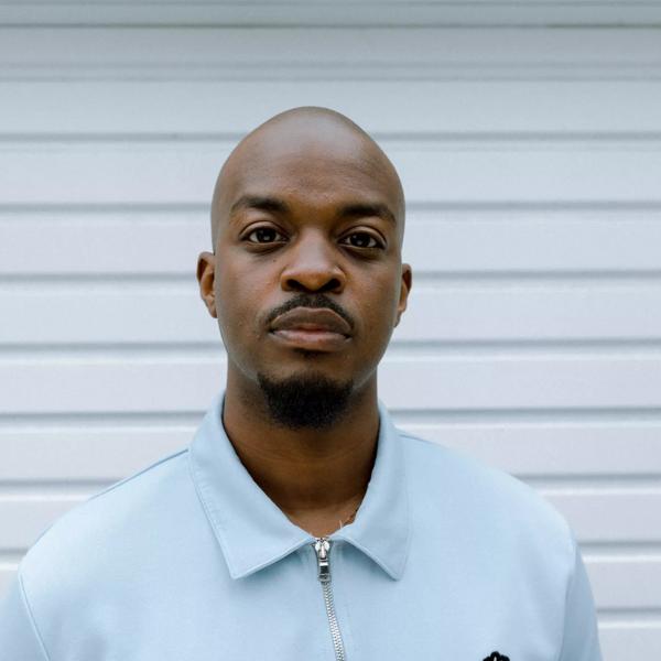 George the Poet wearing  a blue shirt standing in front of a white corrugated background.
