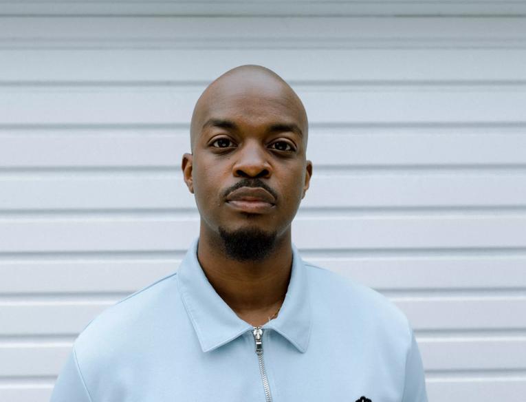 George the Poet wearing  a blue shirt standing in front of a white corrugated background.
