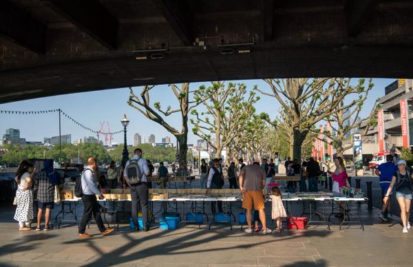 A number of people stand around the tables of the South Bank Book Market under Waterloo Bridge; it is a bright day but the market is in shadow