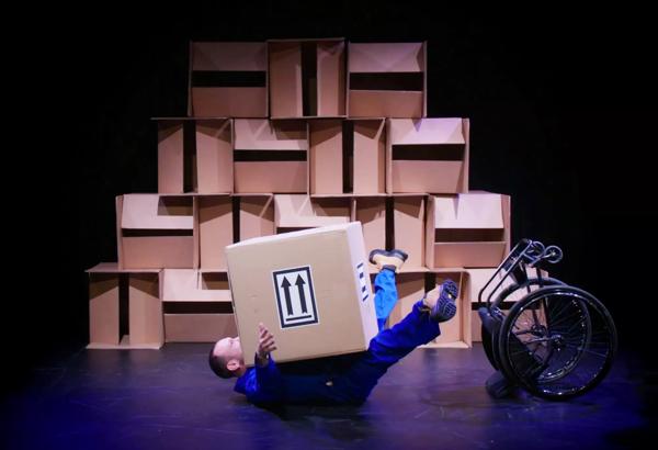 Person wearing a blue boiler suit lying on the floor with their legs in the air and a large cardboard box on top of them and an upturned wheelchair next to them.