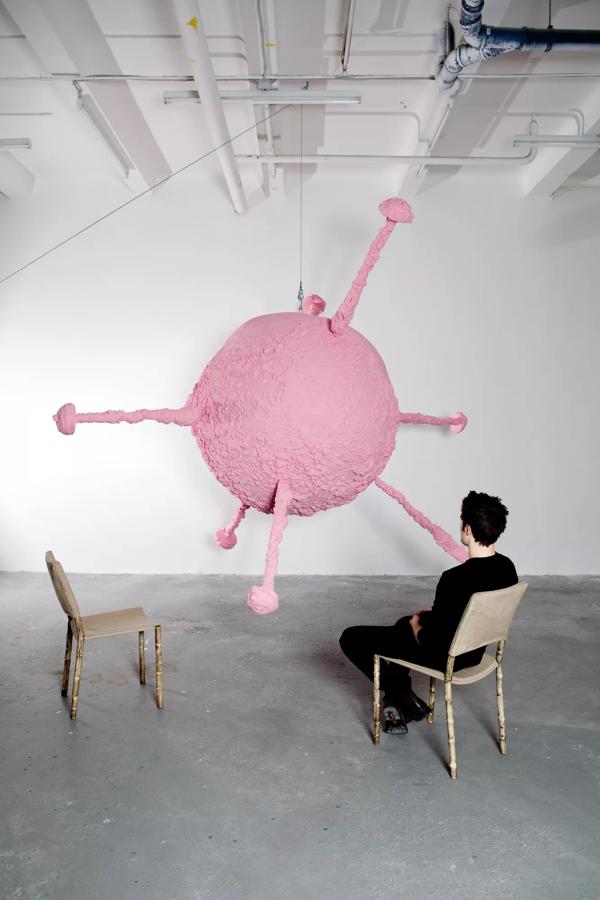 Two wooden chairs sit facing a large pink paper mache balloon like sculpture.