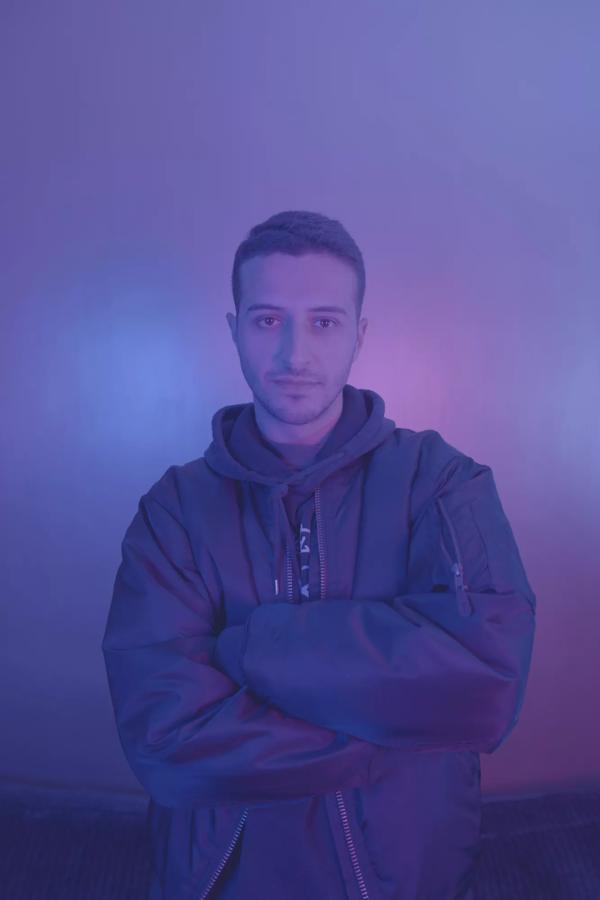 The DJ Kamar stands looking at camera with his arms crossed in a black bomber jacket against a purple lit background. 
