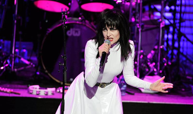 Siouxsie on stage at the 2013 Meltdown Festival, she is wearing a long sleeved white dress and holds a microphone in her right hand, a drumkit is visible behind her