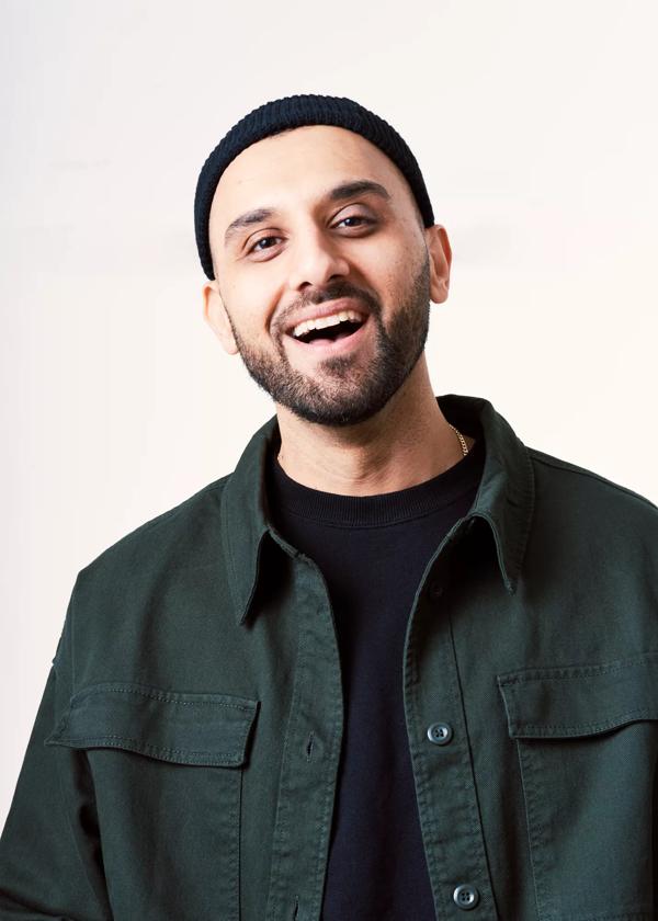 Zohab Zee Khan smiling with mouth open, wearing a black shirt, jacket and hat.