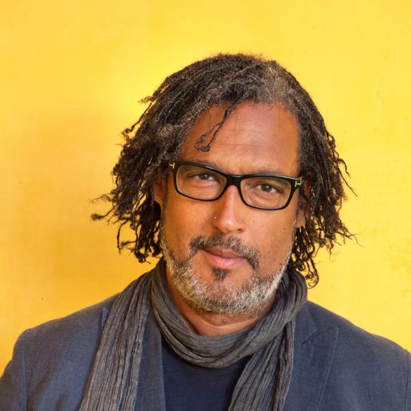 David Olusoga wears a blue scarf and blue jacket in front of a yellow background
