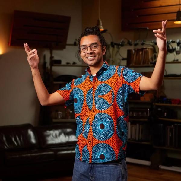The poet Arji Manuelpillai standing in a musical studio, he wears a brightly coloured flowery shirt and is raising his hands in the air