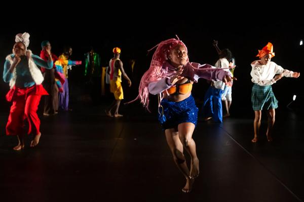A group of dancers perform on a dark stage wearing different colourful outfits