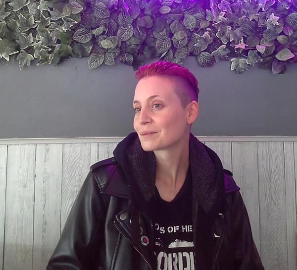 The poet Fran Lock a young White woman with short cropped hair dyed pink. She wears a leather jacket over a black t-shirt and is seated inside against a wall that also features imitation foliage