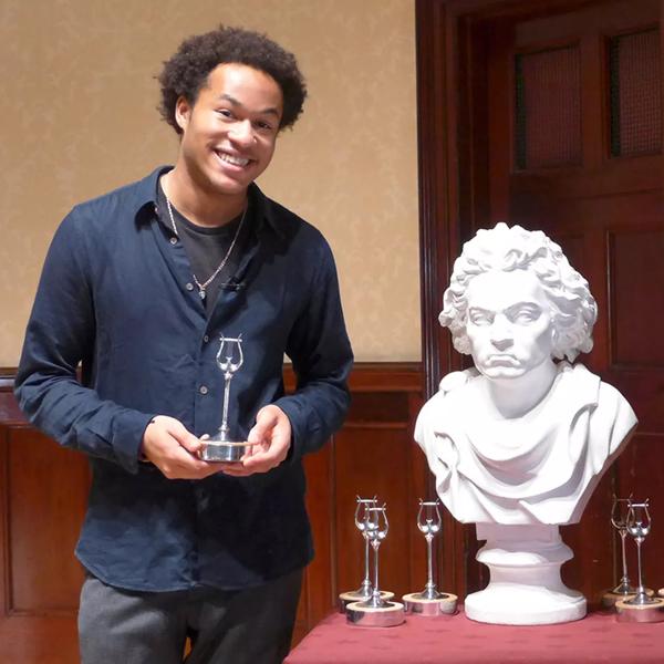 Cellist Sheku Kanneh-Mason holds an award, standing next to a bust of Beethoven