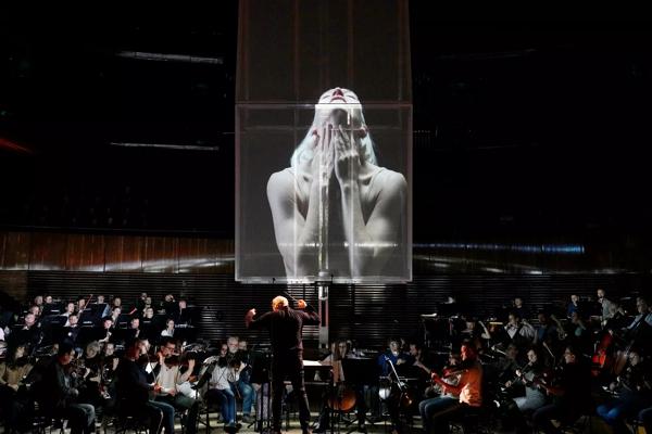 An orchestra with a screen suspended above, displaying dancers.