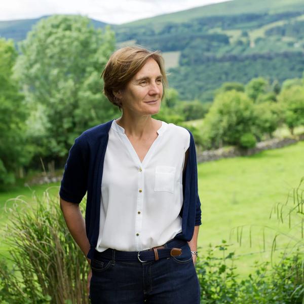 The poet Jane Clarke, a middle aged White woman with short brown hair stands in a field with green hills and countryside behind her