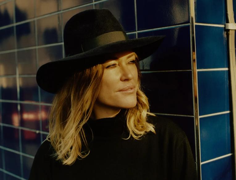 Cerys Matthews stands against a blue tiled wall wearing a black wide-brimmed hat
