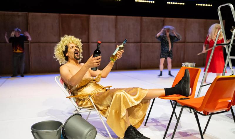 A performer in a gold dress and blonde wig is sat on a chair holding a bottle of wine in each hand 