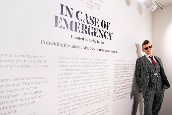 Joelle Taylor, a middle-aged woman who wears a three piece suit and sunglasses with her grey hair styled in a quiff leans against the wall next to a printed notice about the Koestler Arts exhibition, IN CASE OF EMERGENCY, which she has curated