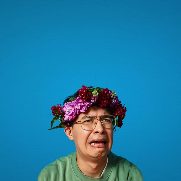Phil Wang hunches against a blue background wearing a crown of purple flowers. He is looking off right of camera and frozen mid-sob. 