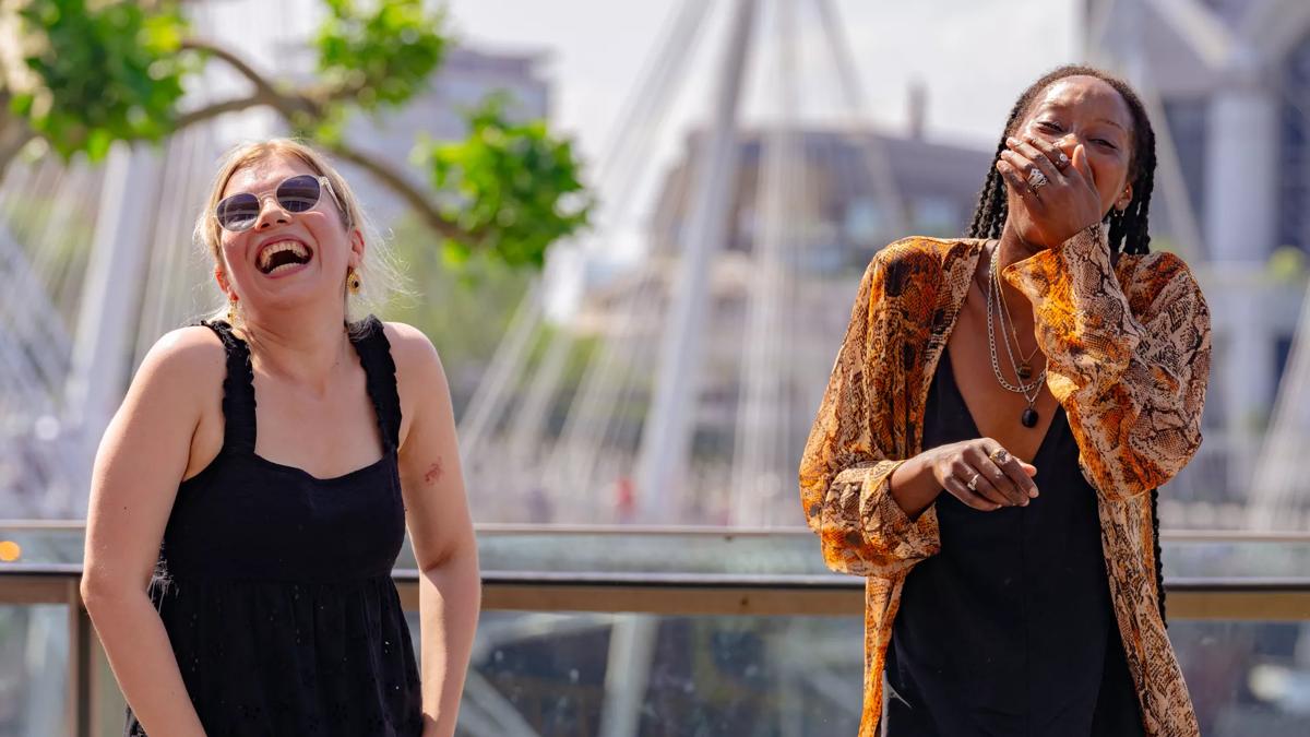 Two women stand laughing in the sunshine.