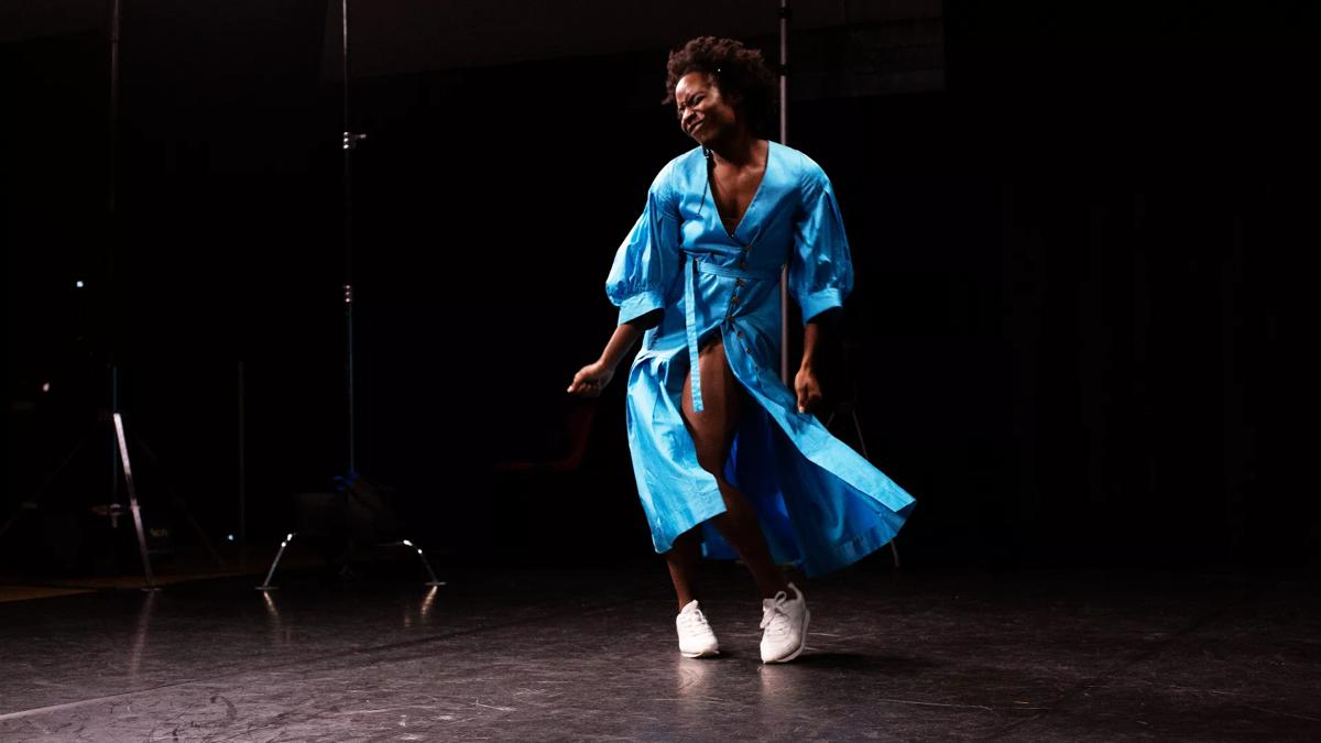 A young Black woman in her sky blue wrap dress dances on a brightly lit stage against a dark backdrop.