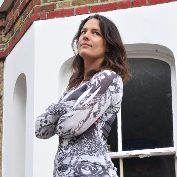 The poet Abigail Parry, a young White woman with long dark hair stands with her arms folded in front of a brick building with a bay window