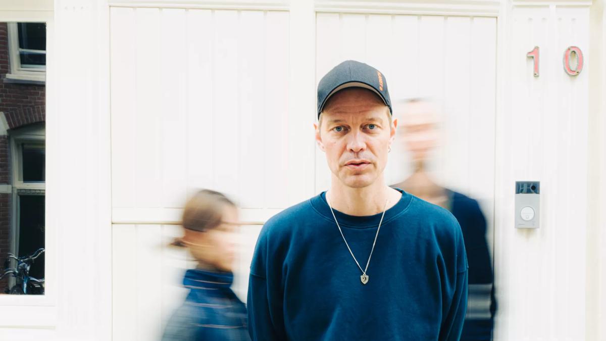 Artist wearing a cap and a blue jumper in front of a blurred background. 