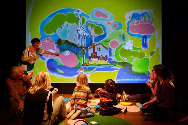 A performer points at a cartoon screen in front of a group of children and adults. 