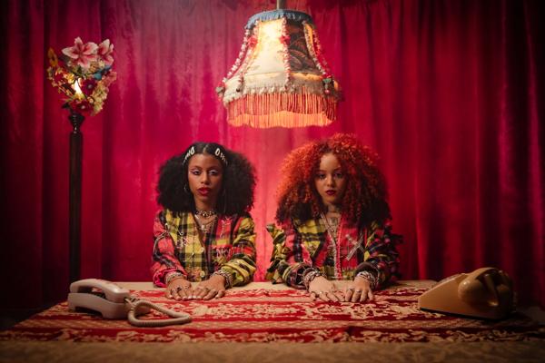 Rock duo Nova Twins at a table with telephones by their side and a ceiling lamp over  their head