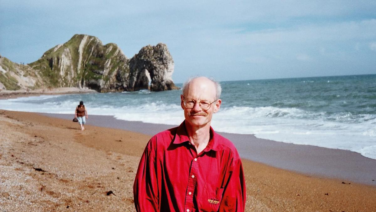 Poet Gavin Selerie wears a red shirt standing on the beach at Durdle Door