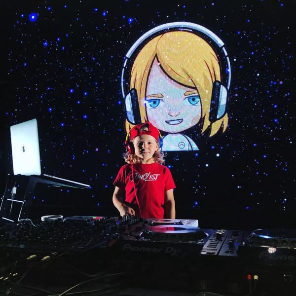 DJ Archie stands behind a set of decks in a red t shirt and red cap.