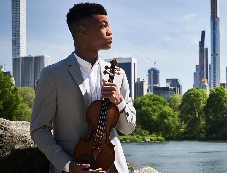 Violinist Randall  Goosby holds violin in park whilst looking into the distance