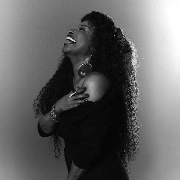 Black and white side profile of the singer Chaka Khan, laughing. She is wearing an over the shoulder black dress. 