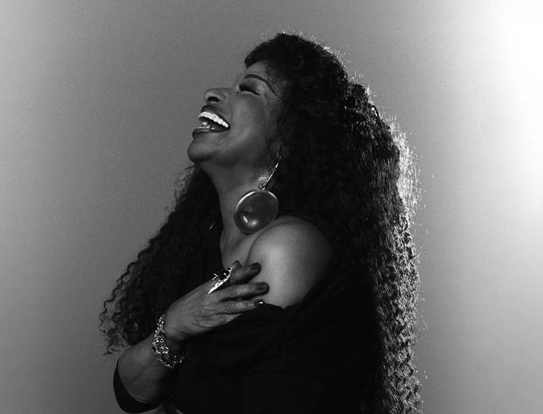 Black and white side profile of the singer Chaka Khan, laughing. She is wearing an over the shoulder black dress. 