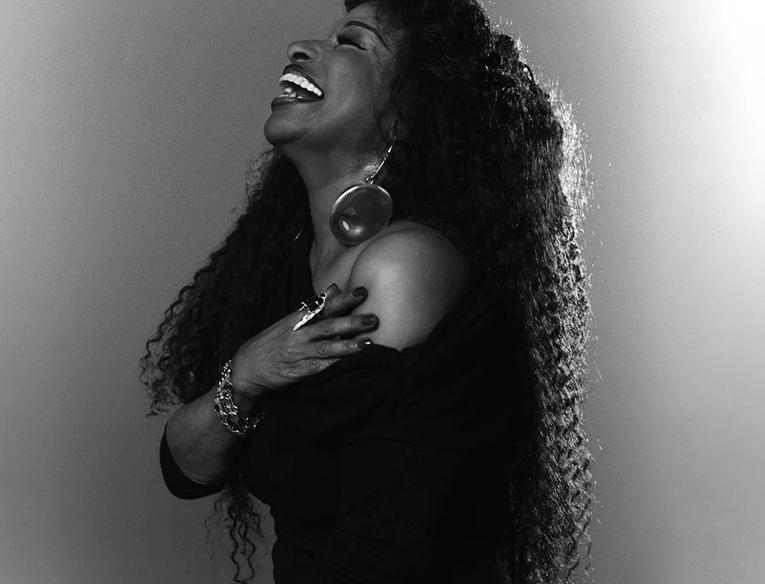 Black and white side profile of the singer Chaka Khan, laughing. She is wearing an over the shoulder black dress. 