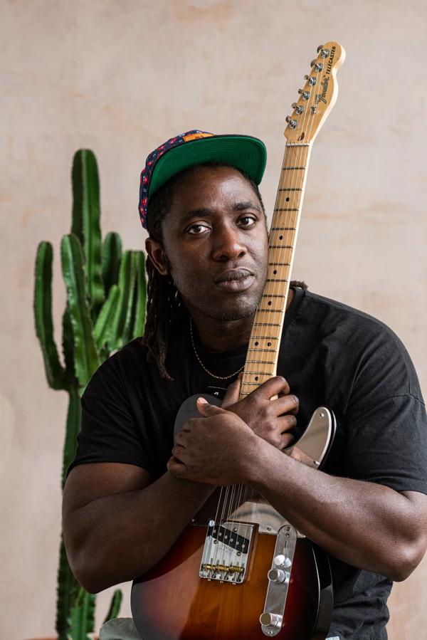 Portrait of musician Kele. He sits in a black t shirt cradling an electric guitar. He has a gold chain on and a cap with a green peak. Behind him stands a cactus. 