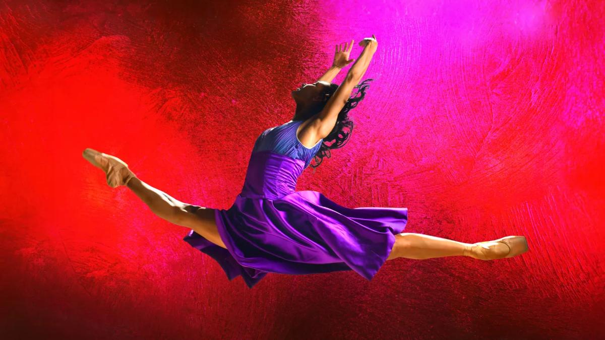 Ballet dancer wearing a purple dress is mid air in a pose against a red and pink background
